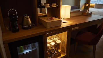 A compact hotel room with a kitchenette, a small refrigerator, a wooden cabinet with glass doors displaying tea sets, a red chair, and a bedside table with a lamp.
