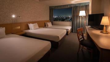 Habitación de hotel con tres camas, un escritorio con televisor y vistas a la ciudad a través de la ventana.
