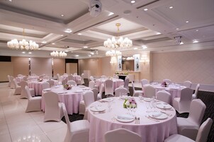 A banquet hall with round tables, white chairs, and chandeliers.