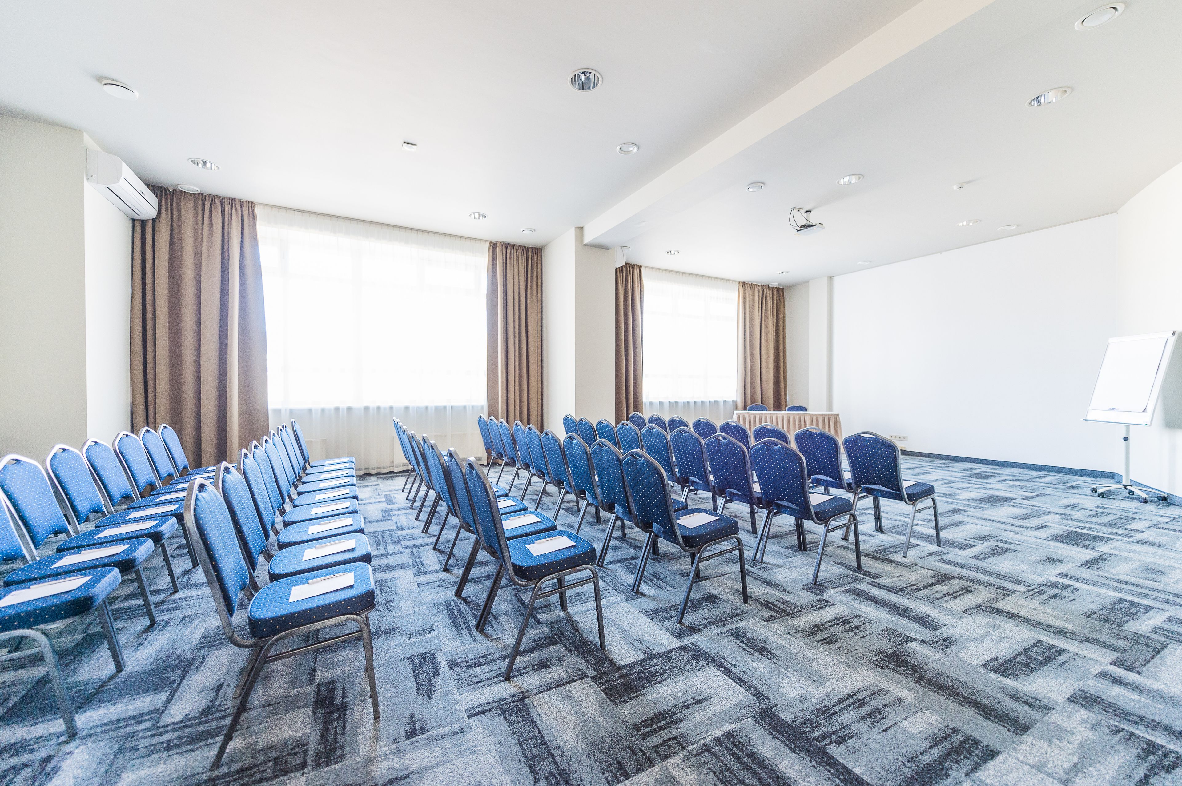 Un salón de conferencias con hileras de sillas azules, suelo alfombrado y amplias ventanas con cortinas.