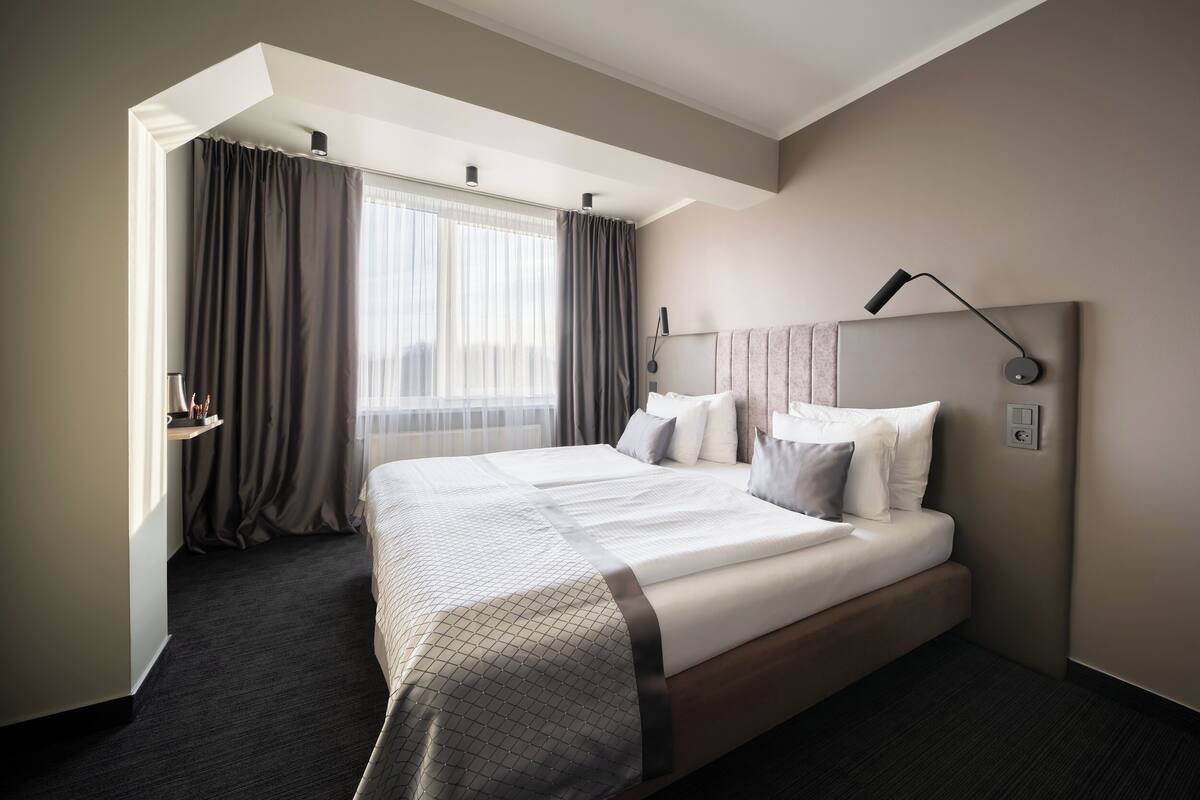 A modern hotel room with a large bed, a bedside table, a lamp, and a window with curtains.