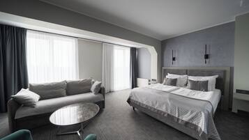Una habitación de hotel moderna con un sofá, una mesita redonda, una cama con cabecera y ventanas grandes con cortinas.