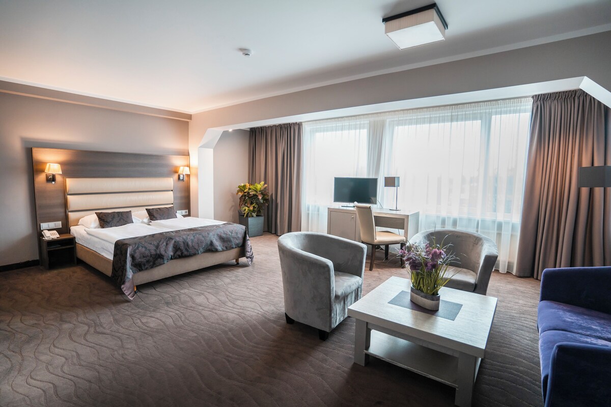 Habitación de hotel moderna con una cama grande, un escritorio, dos sillones y vistas de la ciudad a través de amplios ventanales.