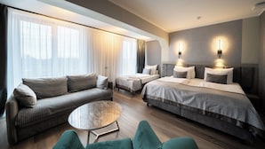 A modern hotel room with a sofa, two single beds, a round coffee table, and a view of the city through large windows. - Bellevue Park Hotel Riga (Riga)