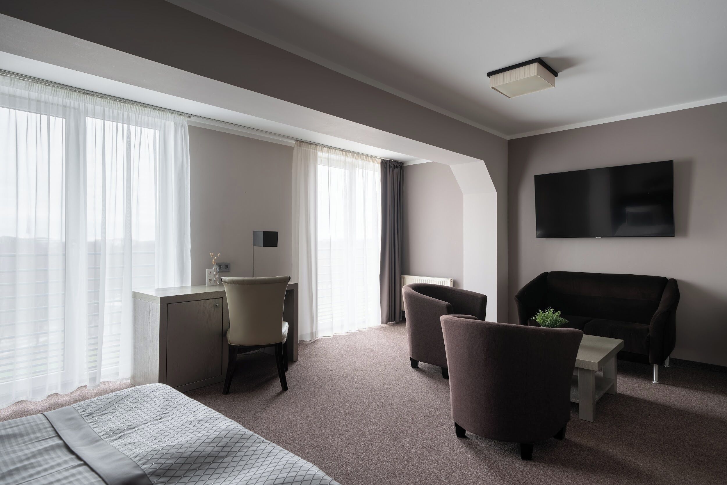 Ett hotellrum med en säng, ett skrivbord, två fåtöljer, en soffa, en tv och stora fönster med tunna, genomskinliga gardiner.