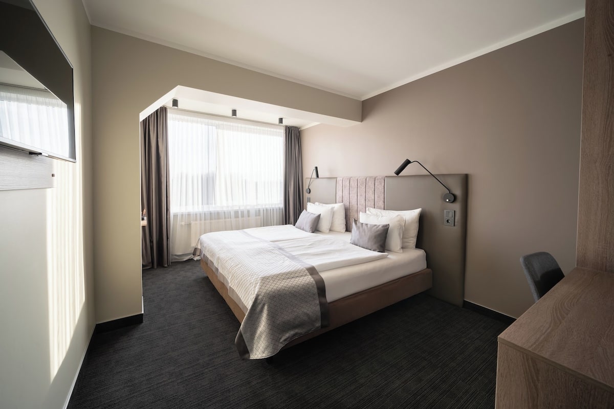A hotel room with a large bed, a desk, a chair, a bedside table, a lamp, and a window with curtains.