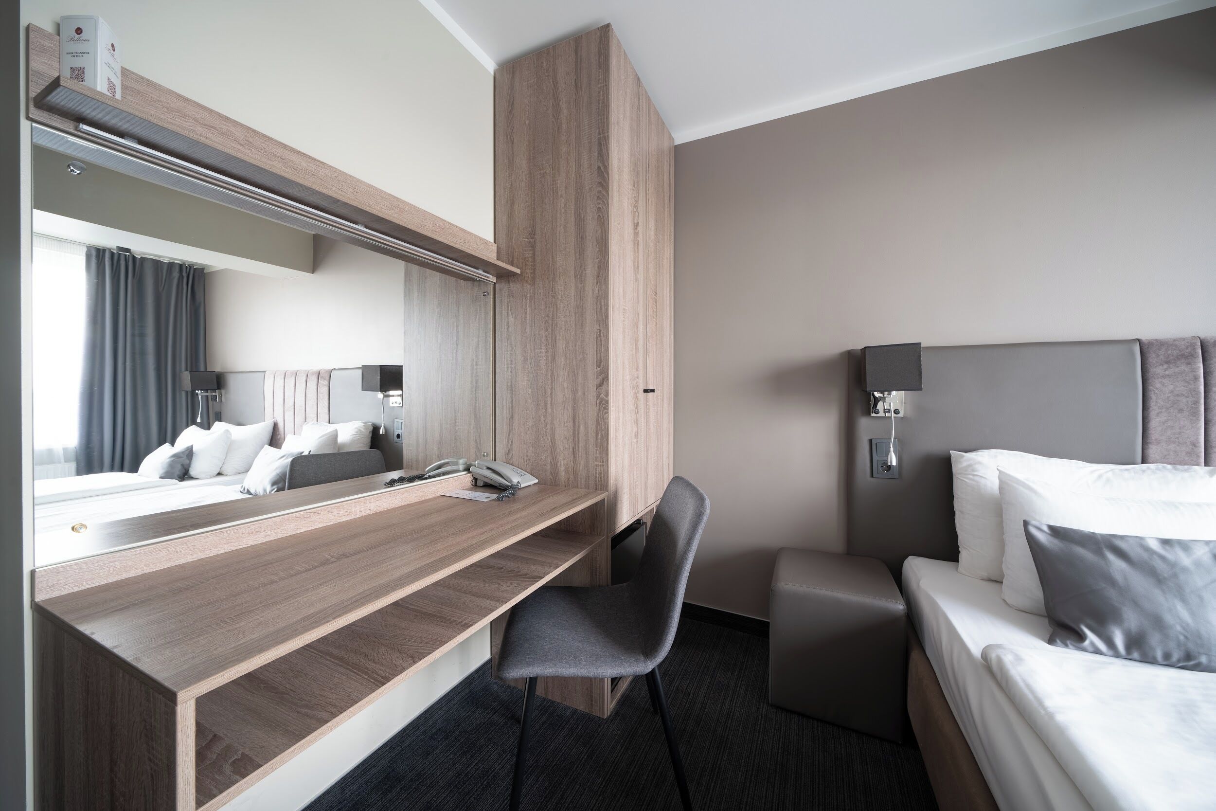 A modern hotel room with a wooden desk, a comfortable chair, a bed with white linens, and a large mirror.