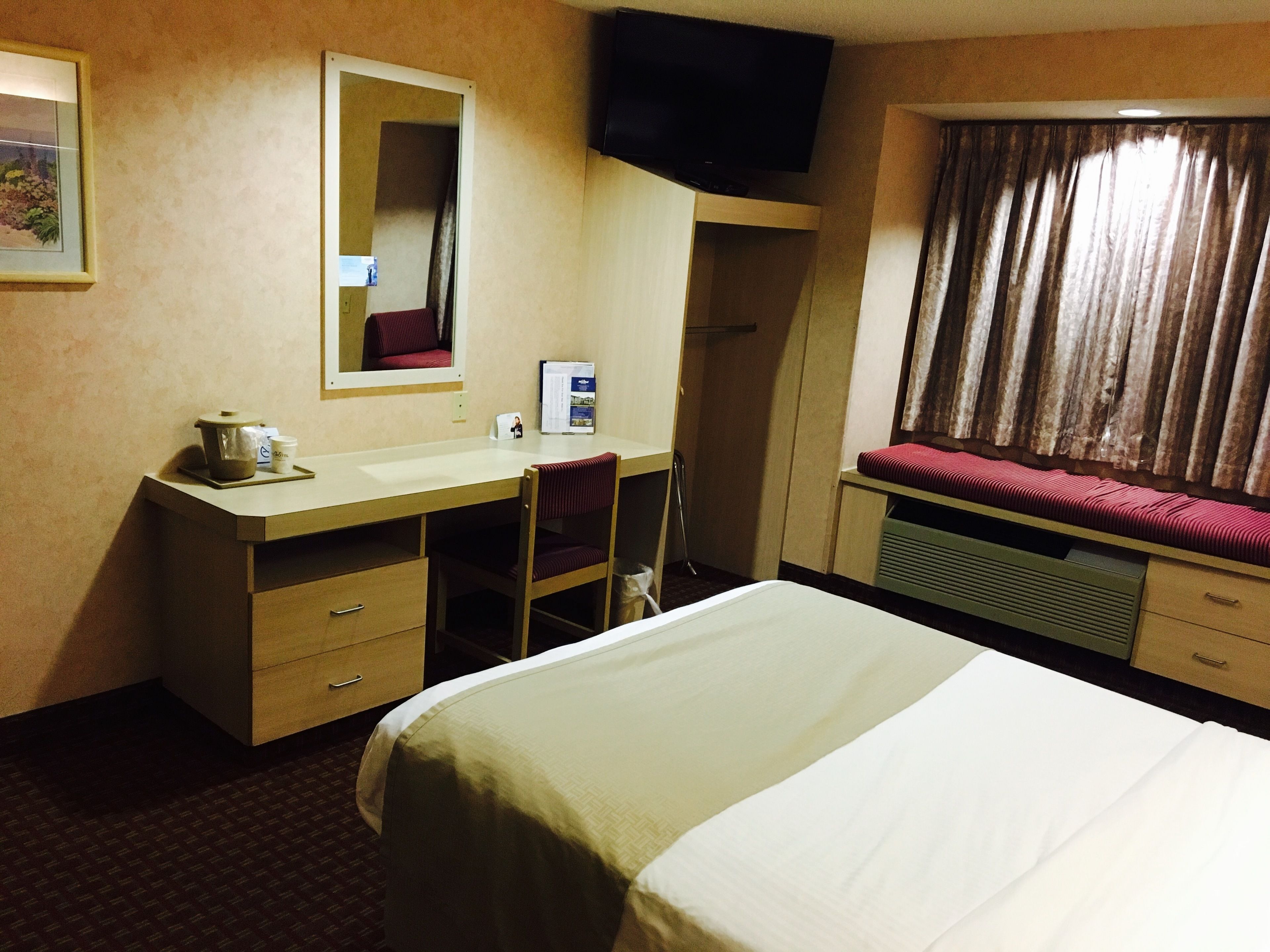 A hotel room with a bed, a desk with a chair, a television mounted on the wall, and a window with curtains.