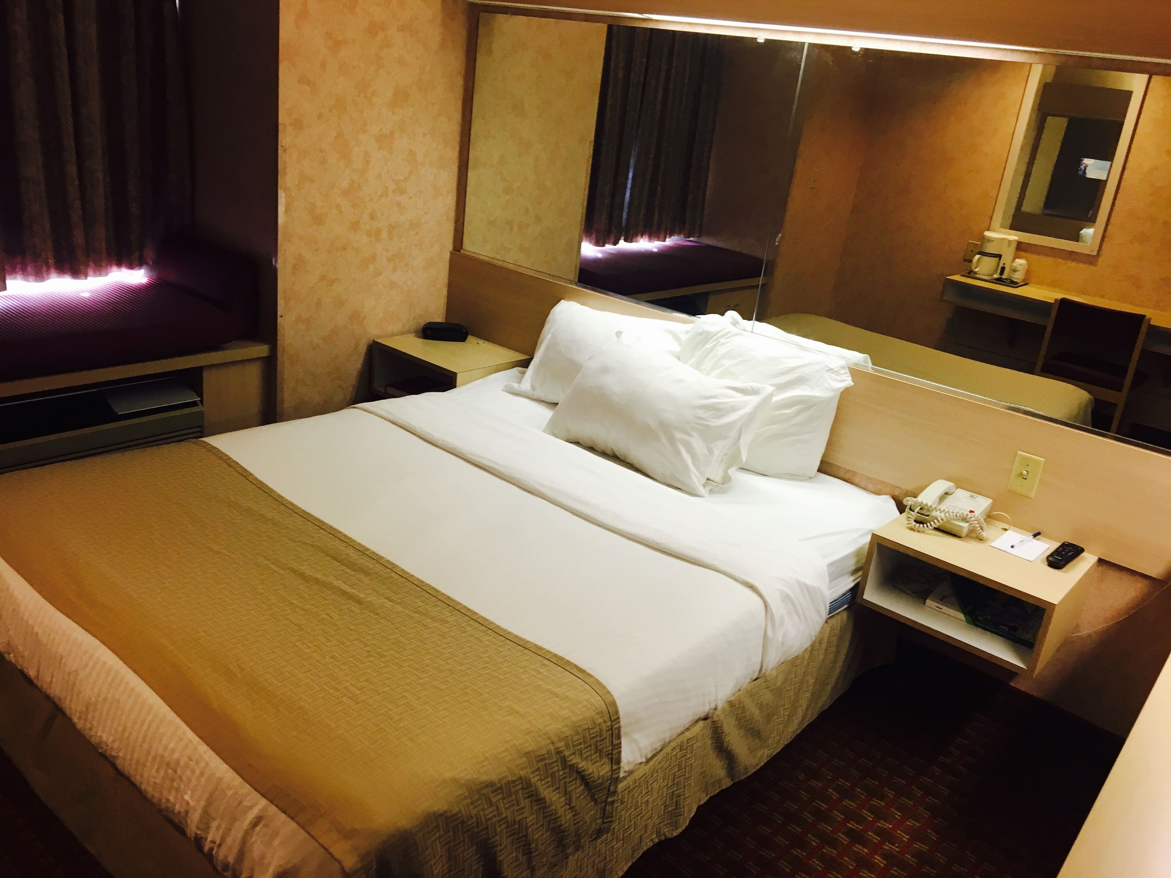 A hotel room with a bed, a bedside table with a phone and a lamp, a mirror, and a window with curtains.
