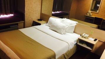 A hotel room with a bed, a bedside table with a phone and a lamp, a mirror, and a window with curtains.