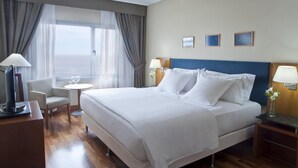 Ein Hotelzimmer mit einem großen Bett, einem Schreibtisch mit Lampe, einem Sessel, einem kleinen Tisch mit Blumenvase und Meerblick durch das Fenster.