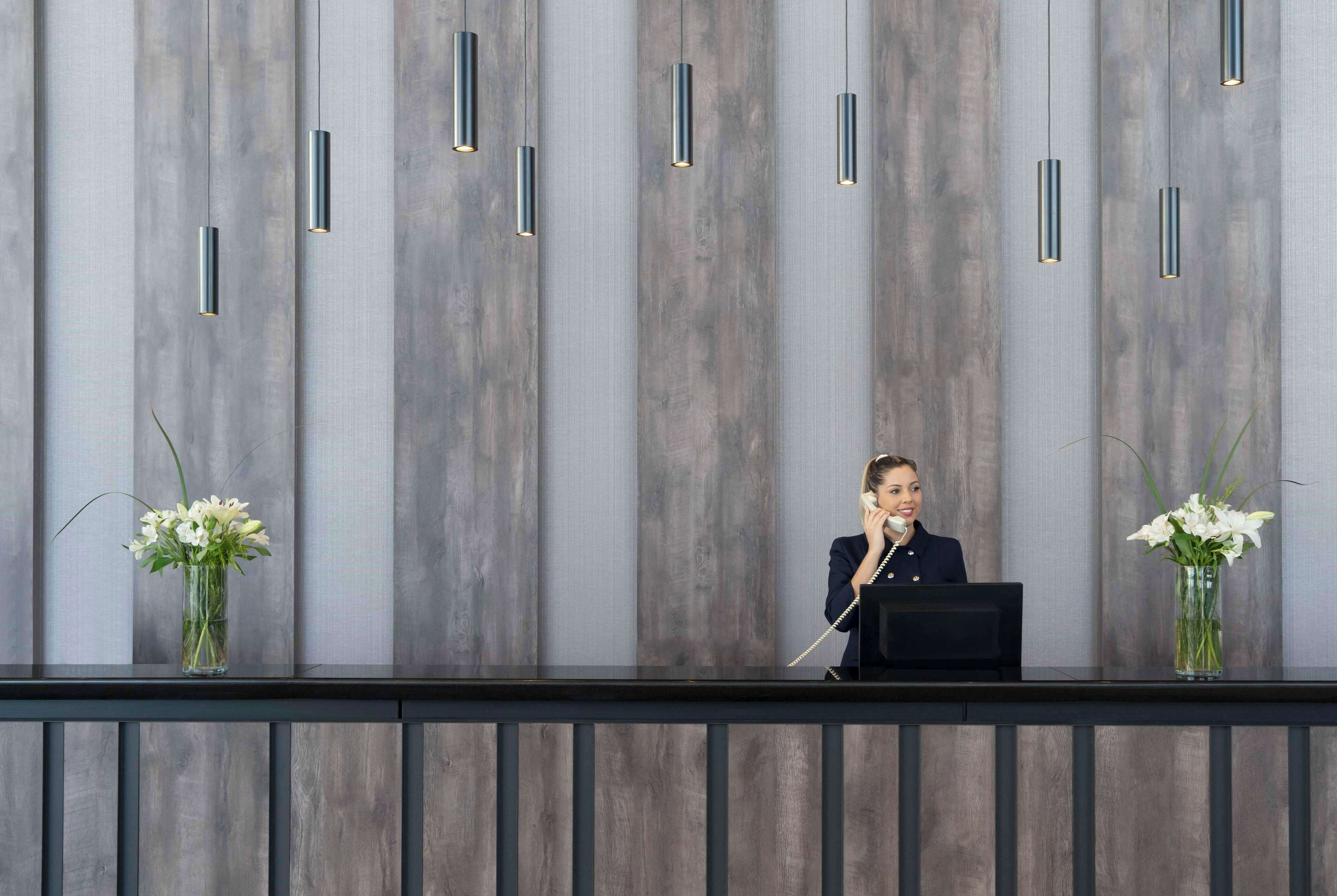 Un área de recepción con diseño moderno, que incluye un panel de madera, un mostrador de recepción y dos jarrones con flores blancas.