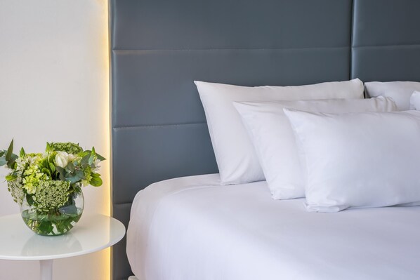 Una cama bien hecha con ropa blanca y almohadas, una pequeña mesa redonda con un jarrón de flores y un cabecero tapizado en gris.