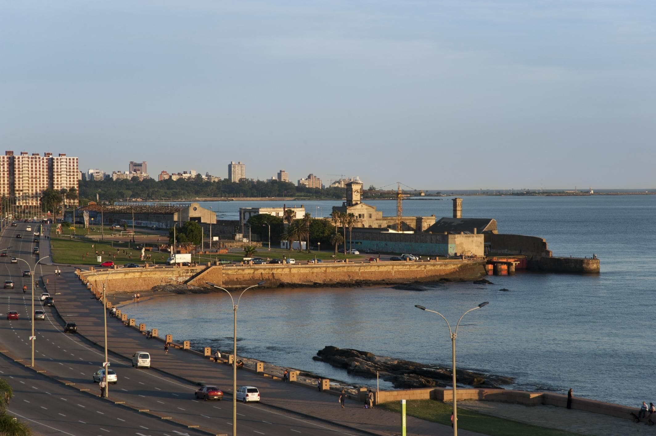 Un paisaje urbano costero con una fortaleza destacada, una calle concurrida y una vista despejada del mar.