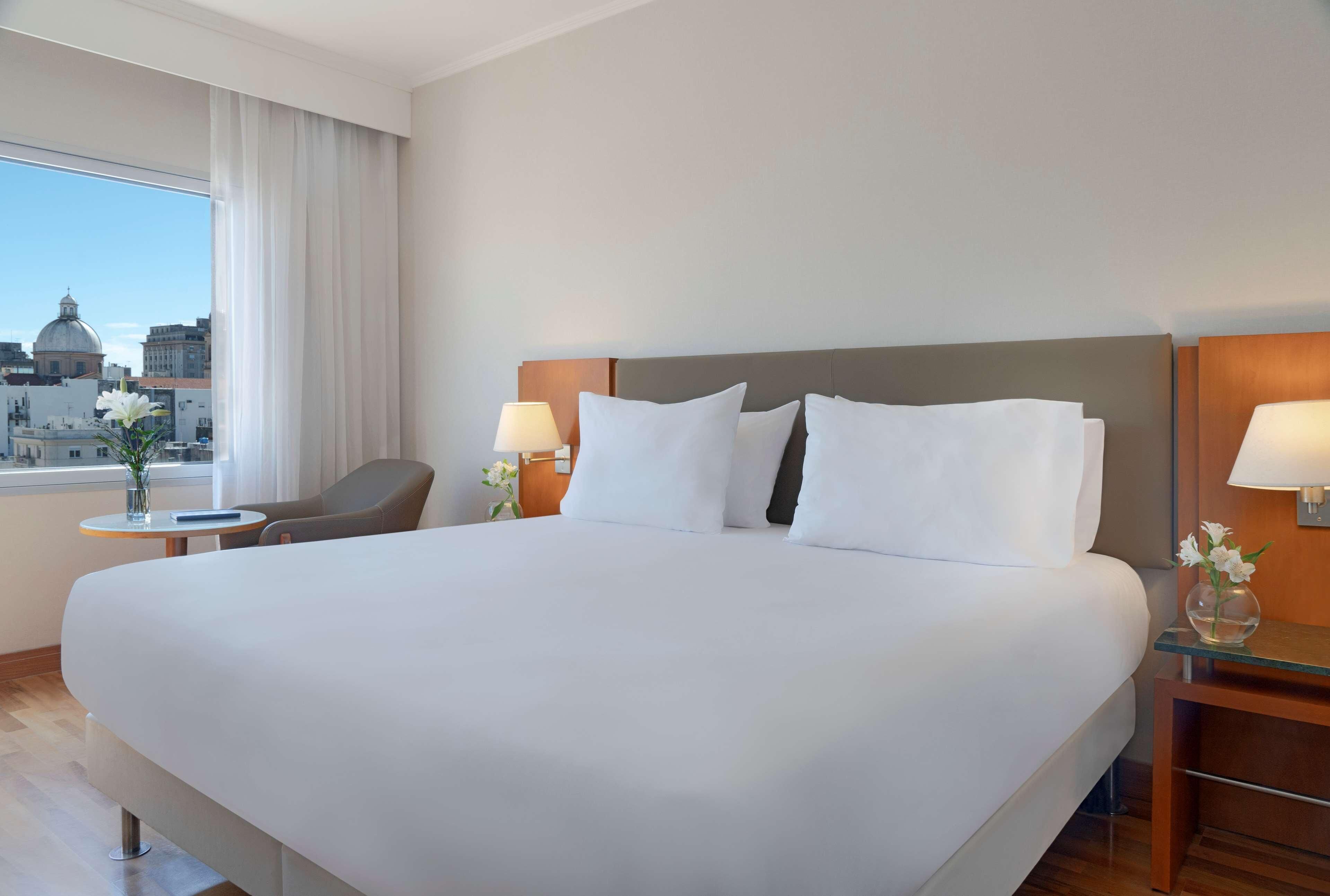 A hotel room with a large bed, two bedside tables with lamps, a chair, a small table with a vase, and a view of the cityscape.