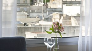 Ein Hotelzimmer mit Blick auf eine Stadtlandschaft, ein Tisch mit einem Buch und einer Blumenvase sowie ein Stuhl.
