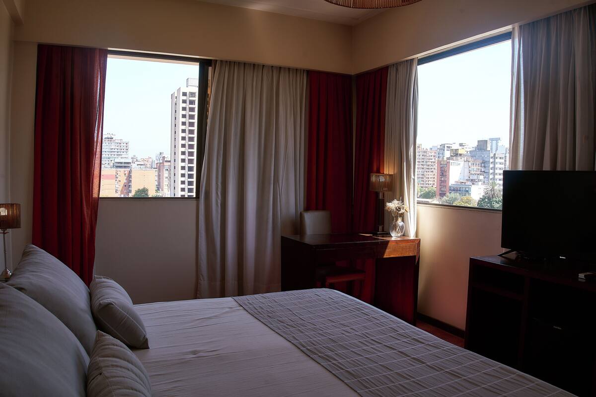 Ein Hotelzimmer mit einem großen Bett, einem Schreibtisch mit Stuhl, einem Fernseher und einem Fenster mit Vorhängen.
