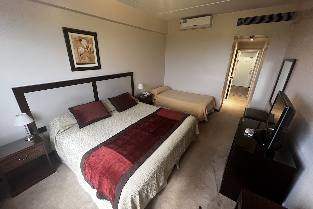Ein Hotelzimmer mit Bett, Nachttischen, einer Lampe, einem Fernseher und einem gerahmten Bild an der Wand.