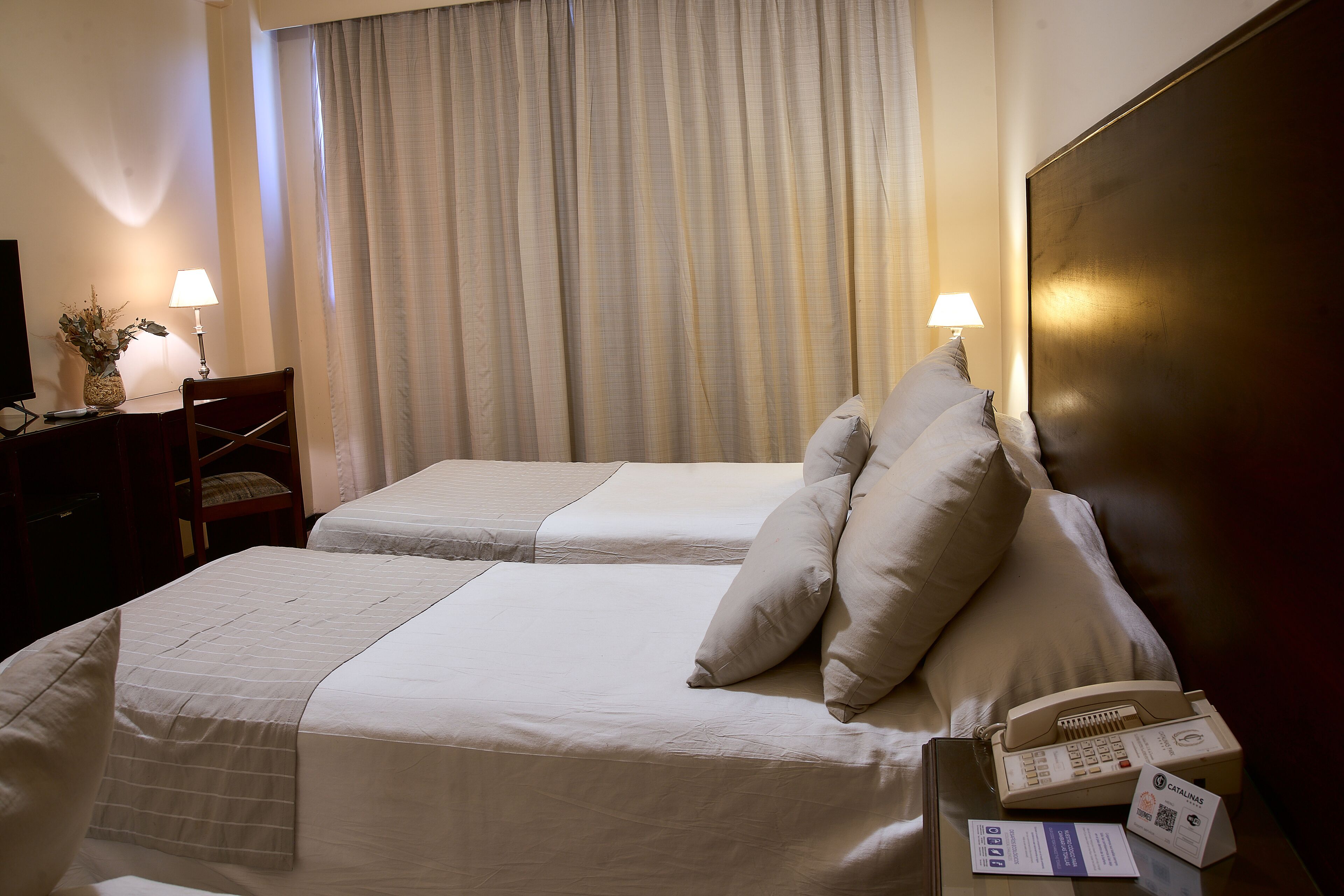 A hotel room with two beds, a desk with a lamp, a telephone, and a vase with flowers.