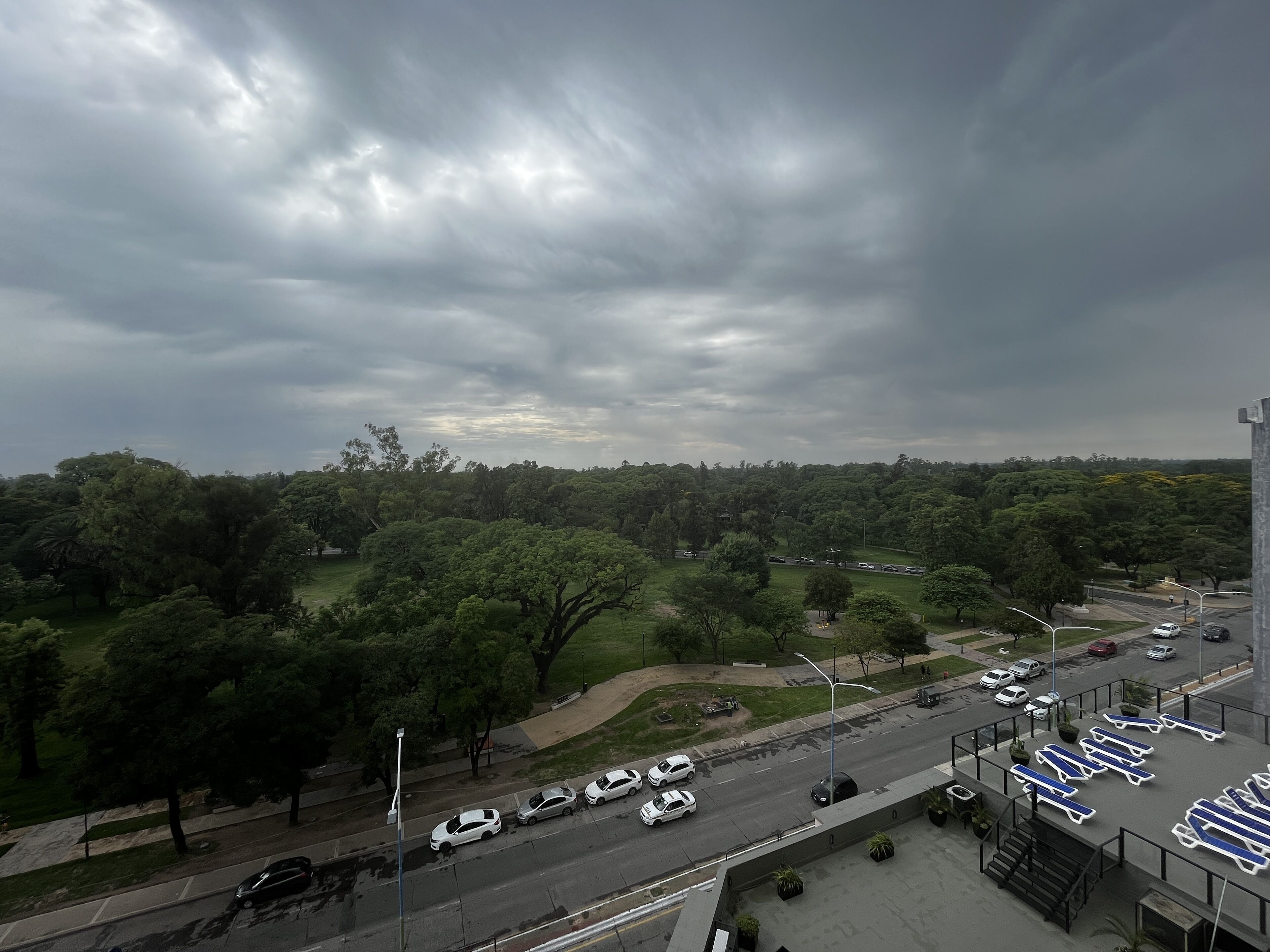Vista aérea de un estacionamiento con varios coches, un parque verde al fondo y un cielo nublado.