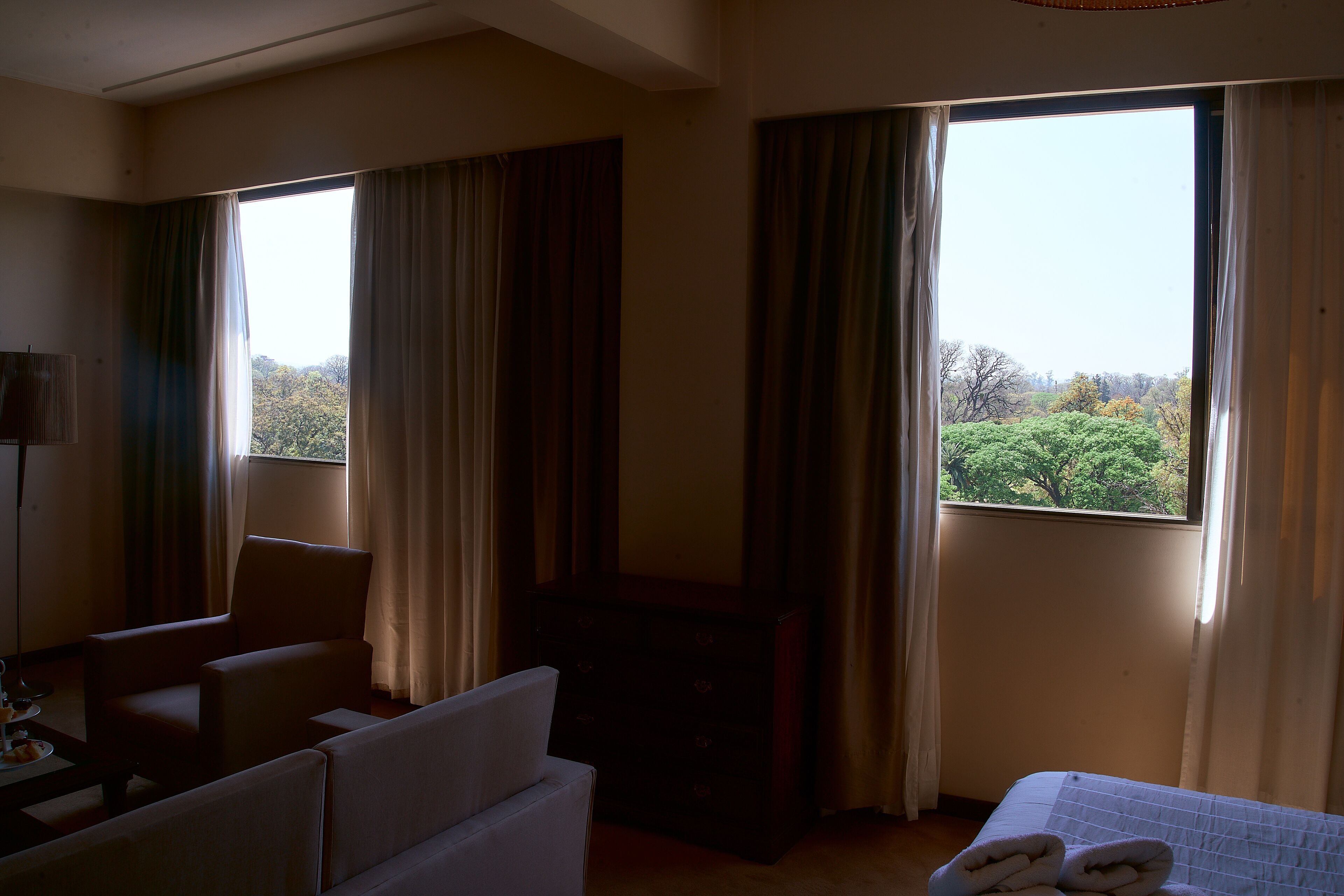 A hotel room with a bed, a chair, a small table, and a view of trees through the window.