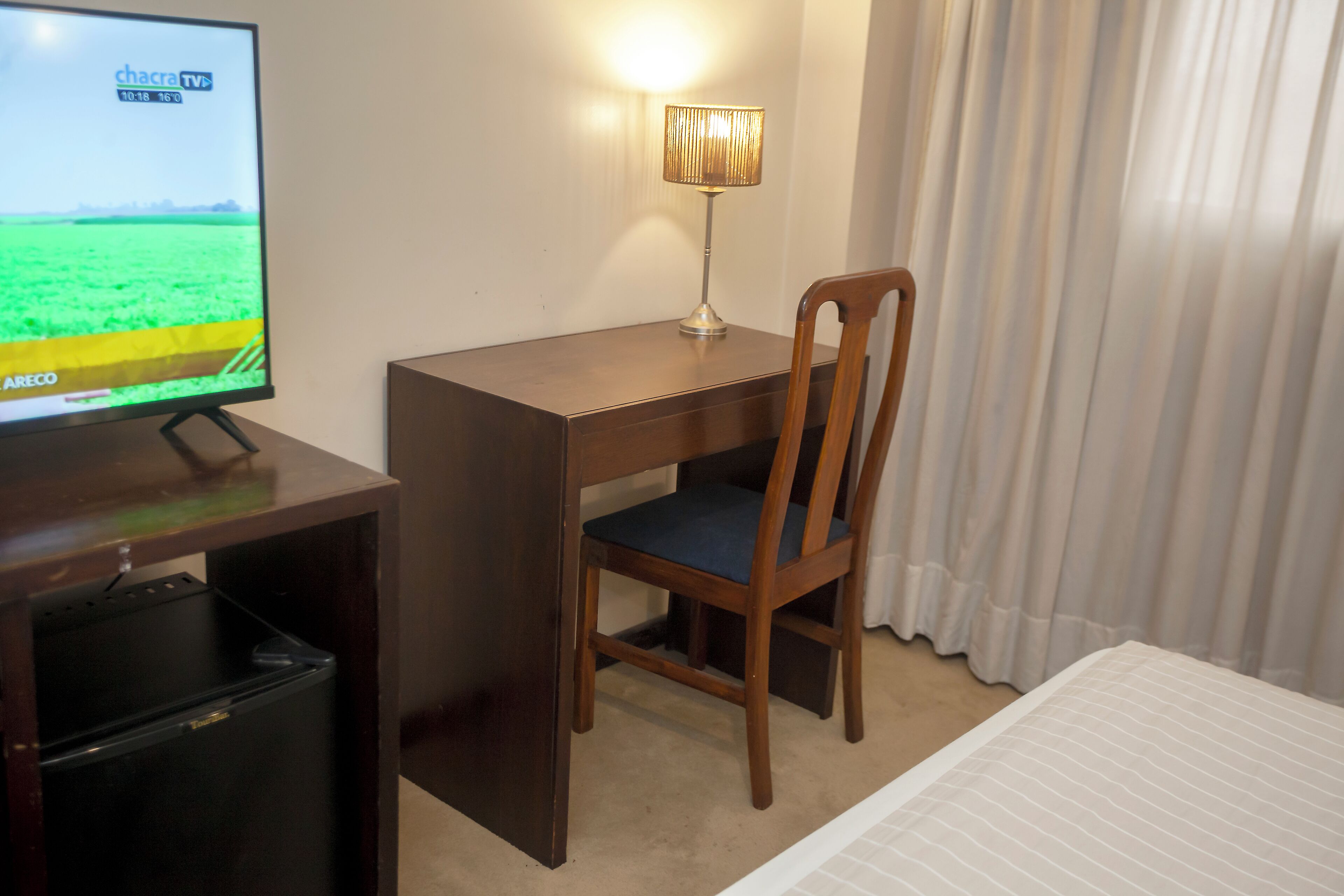 A hotel room with a wooden desk, a chair, a television, and a lamp.