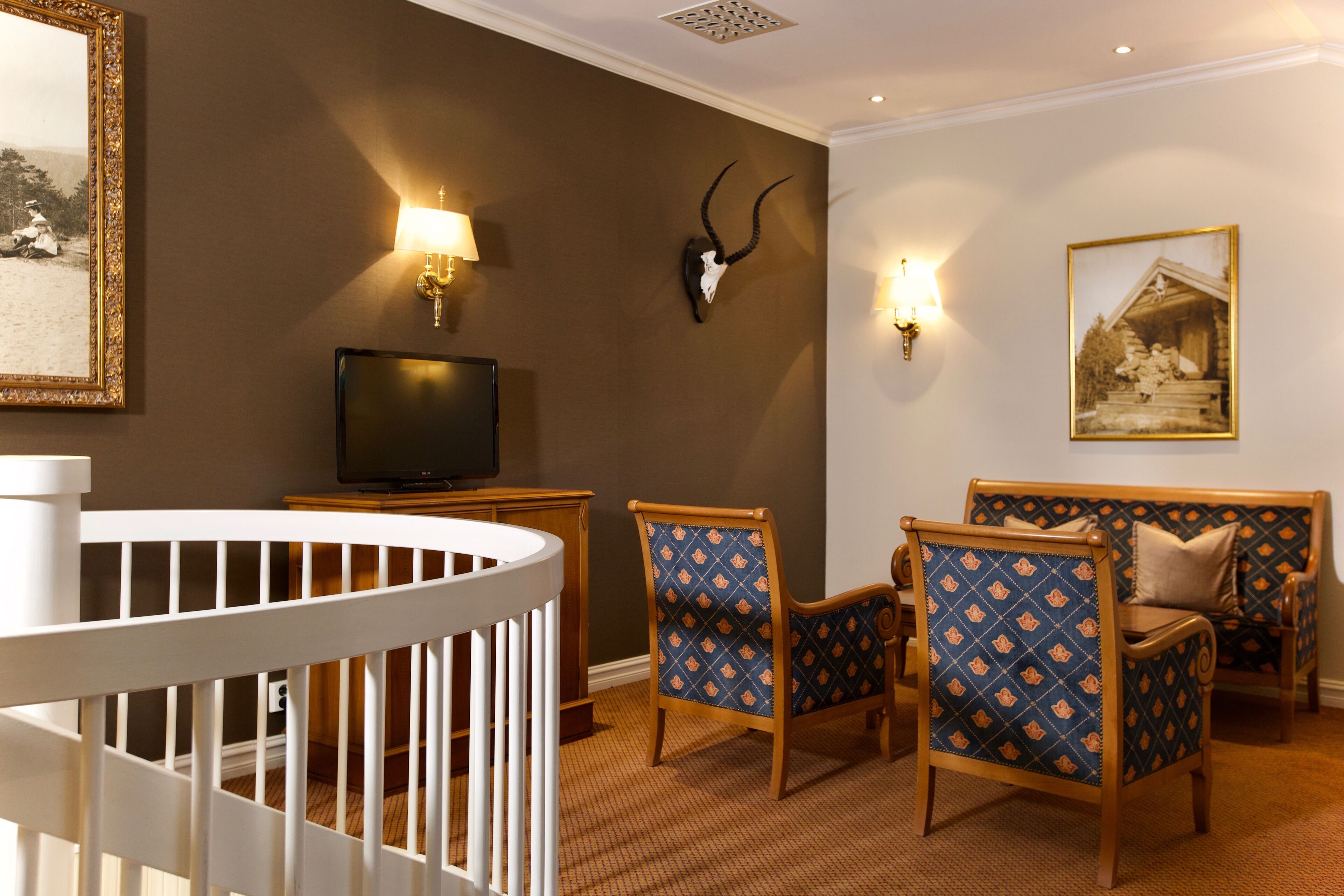 Une pièce avec un berceau blanc, deux fauteuils à motifs, un téléviseur, des lampes fixées au mur et un crâne d’animal accroché au mur.