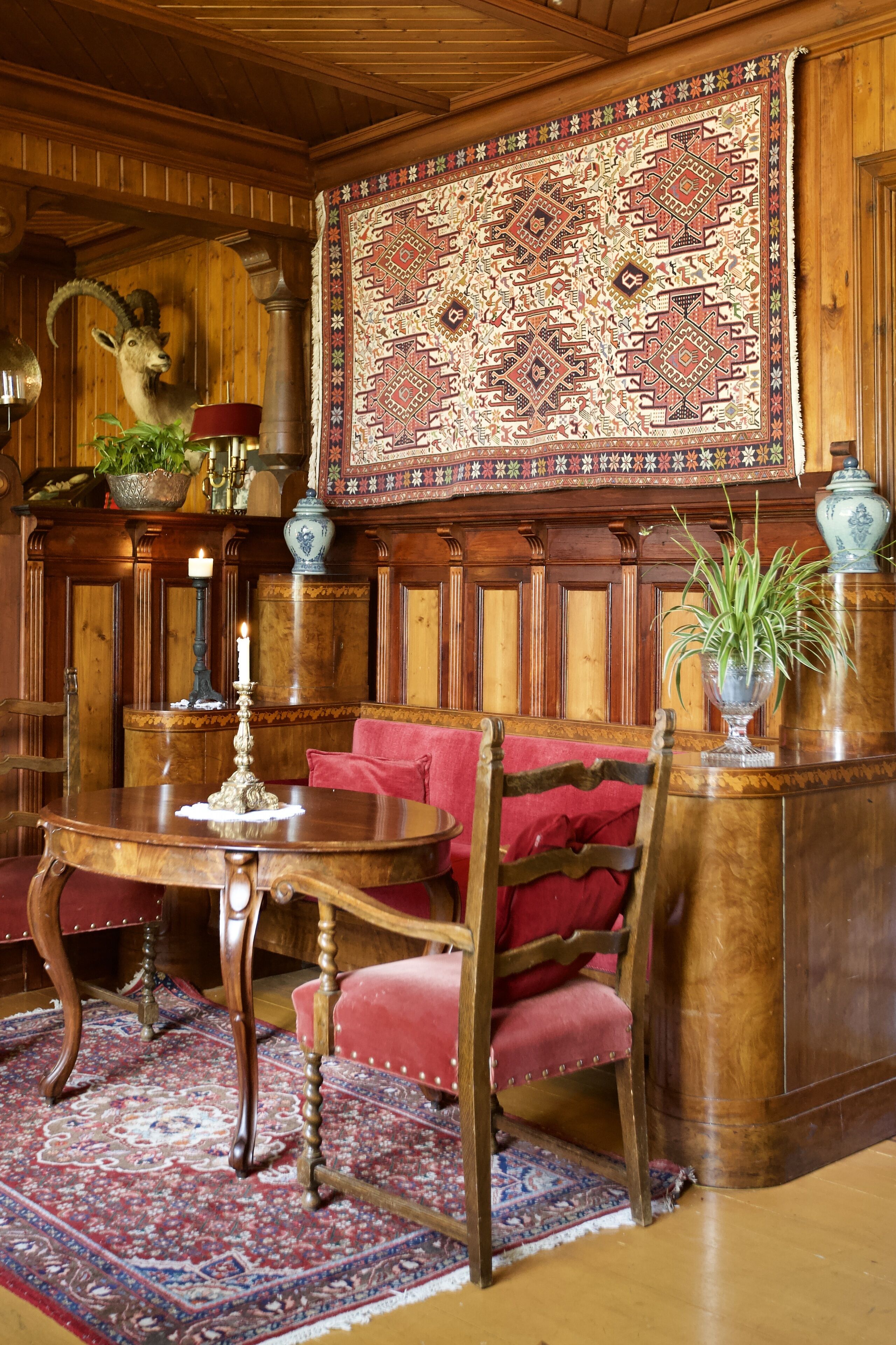 Quarto tradicional com painéis de madeira, um tapete grande com padrão, um sofá vermelho, uma mesa de madeira com uma vela e uma cabeça de animal decorativa.