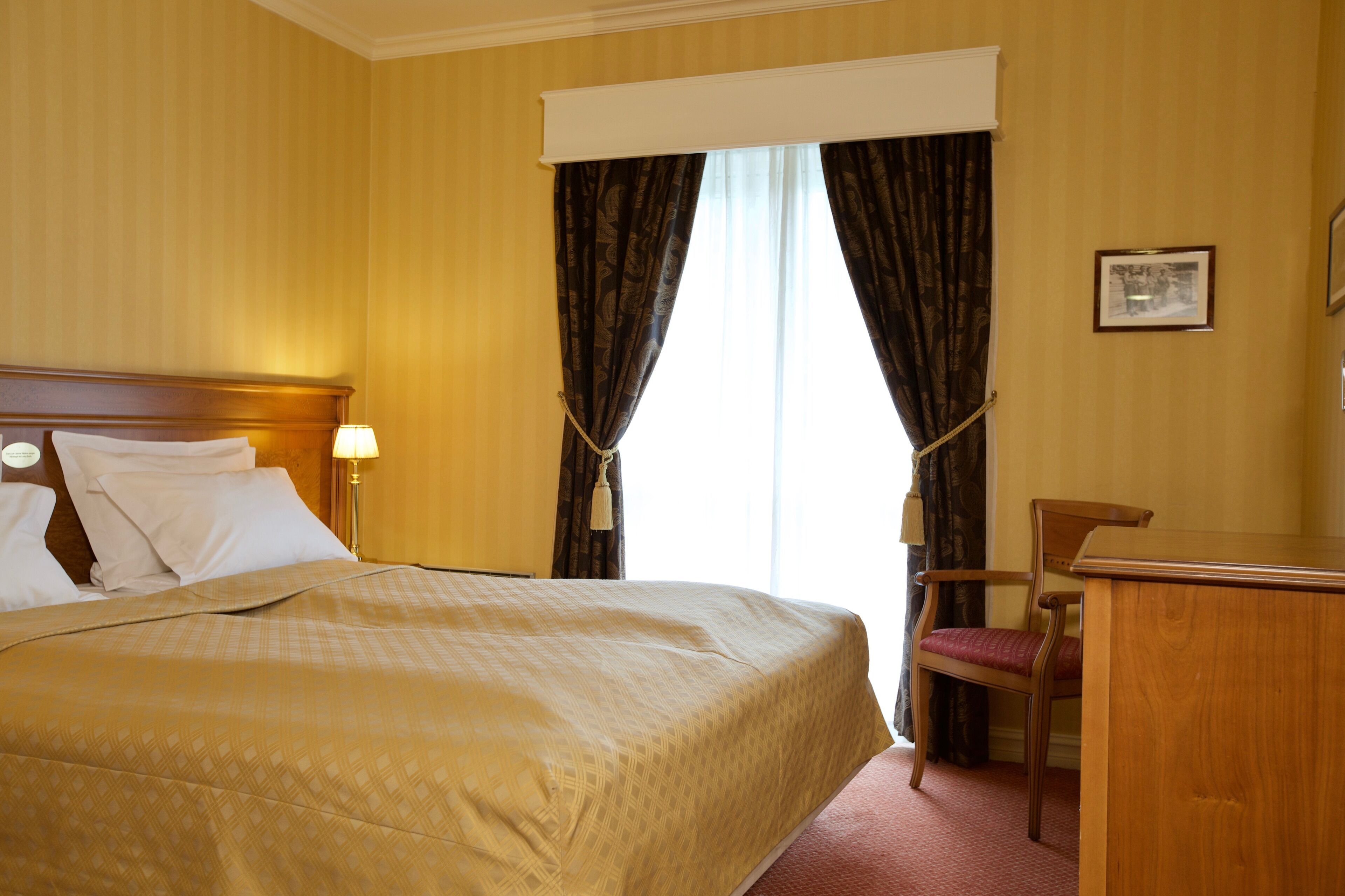 Quarto de hotel com uma cama grande, uma escrivaninha de madeira, uma cadeira e uma janela com cortinas.