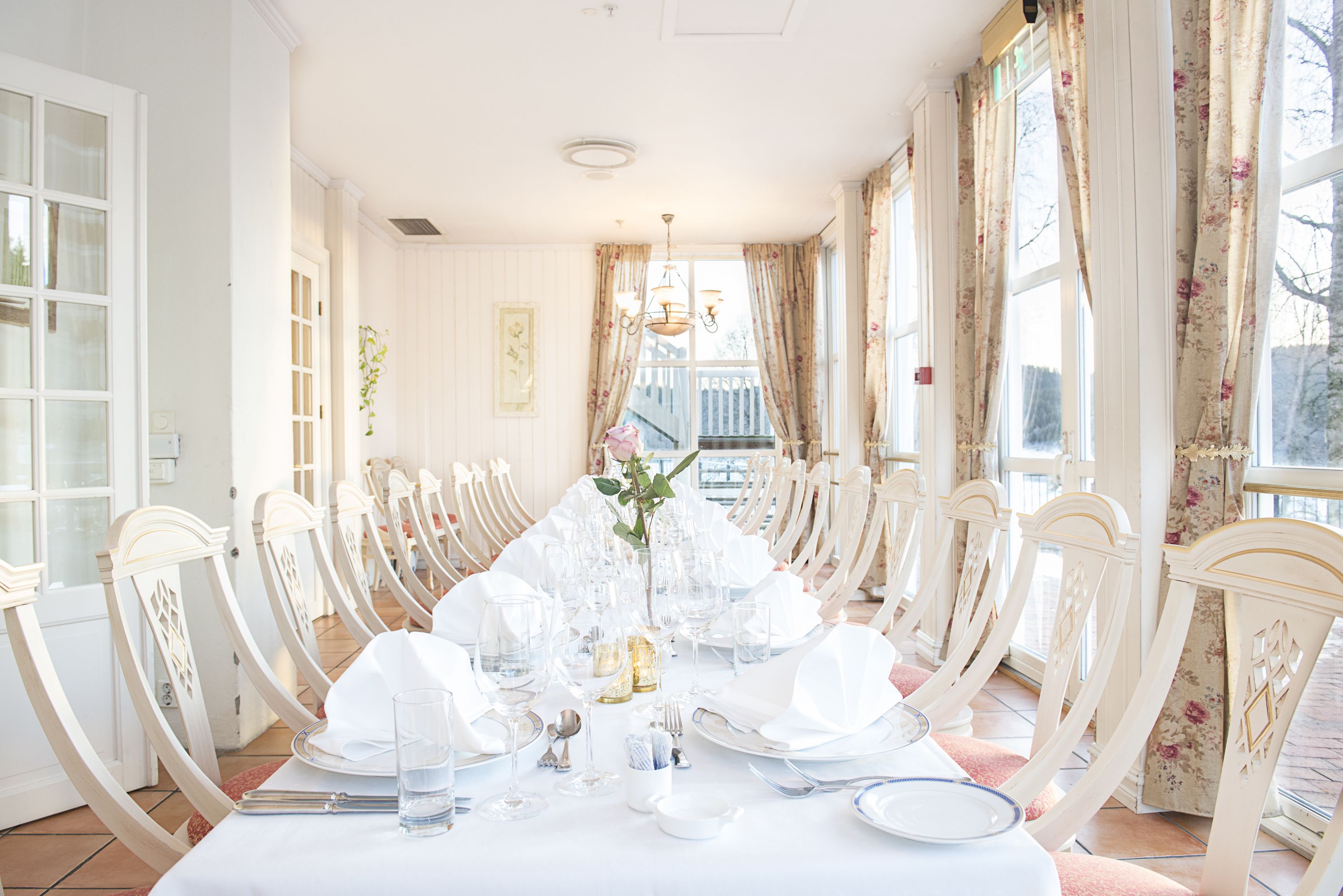 Un comedor con una larga mesa dispuesta para una comida, rodeada de sillas blancas, y vista a una terraza de madera.