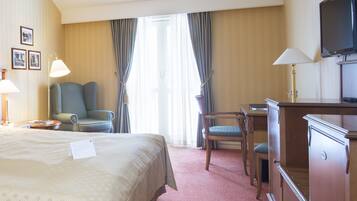 Una habitación de hotel con cama, dos sillones, un escritorio, televisor y ventana con cortinas.