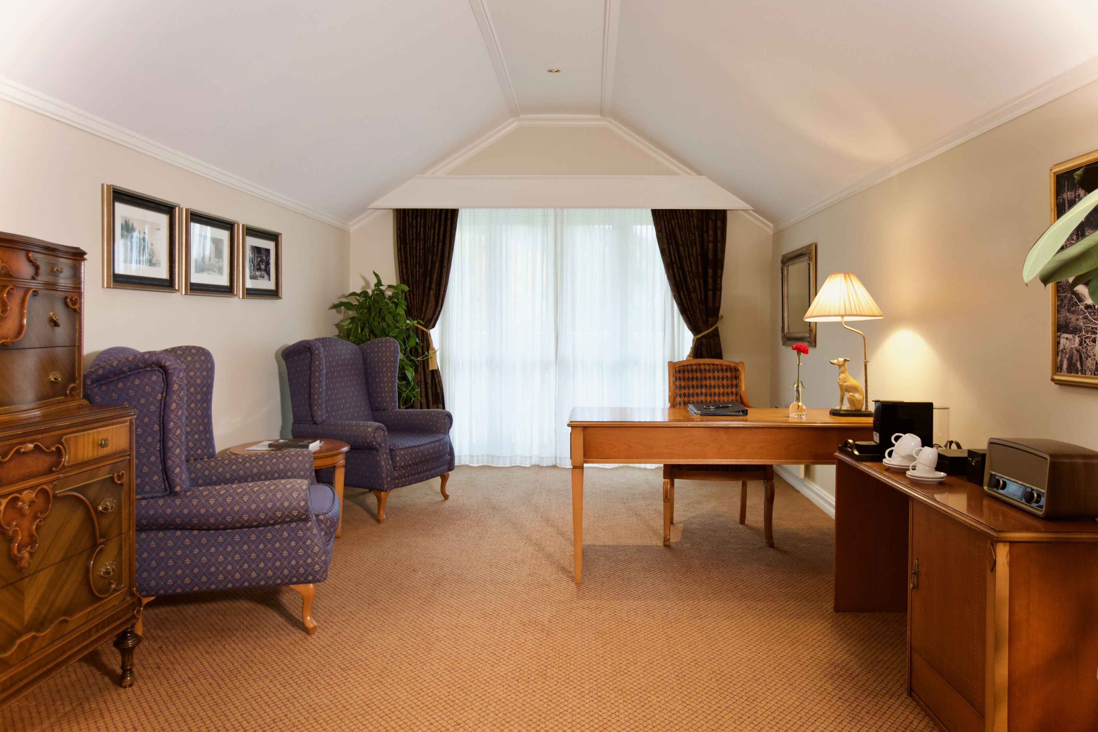 A hotel room with a wooden desk, two armchairs, a dresser, a lamp, and a radio.