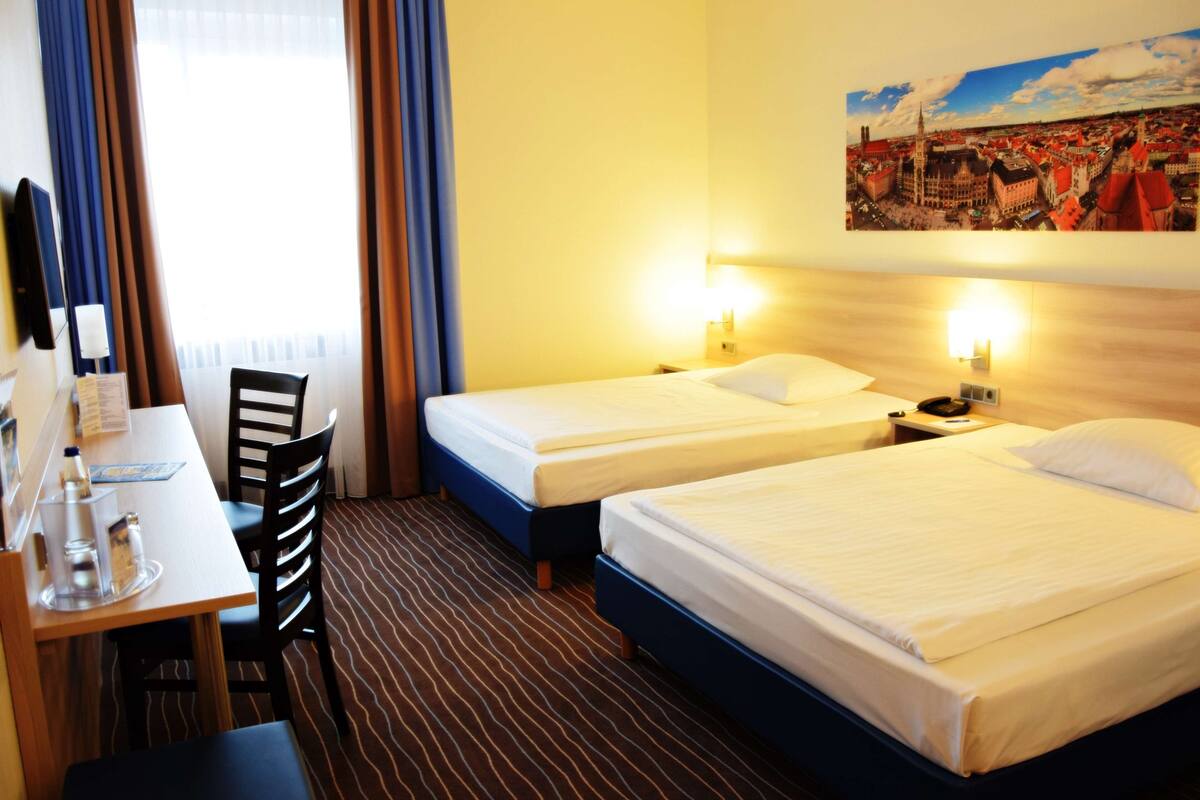 Una habitación de hotel con dos camas, un escritorio, sillas y una pintura de un paisaje urbano en la pared.