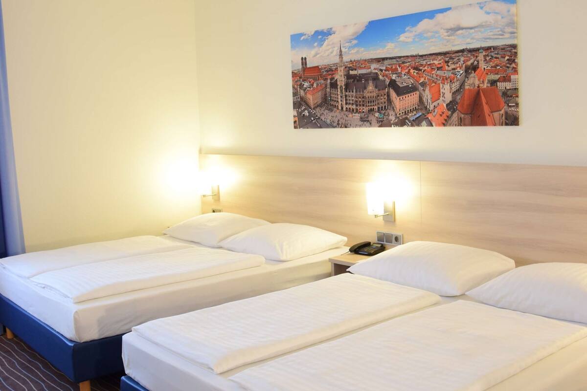 Habitación de hotel con dos camas, un cuadro de un paisaje urbano en la pared y una mesita de noche con lámpara.