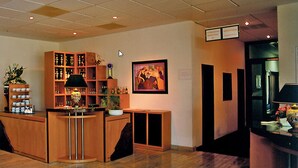 Um hall de hotel com uma mesa de recepção de madeira, prateleiras com diversos objetos, um quadro emoldurado e um vaso de planta.