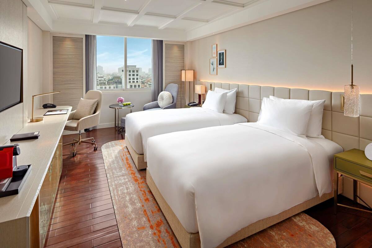 Ein Hotelzimmer mit zwei Betten, einem Schreibtisch, einem Stuhl, einem Fernseher und einem Fenster mit Blick auf die Stadt.