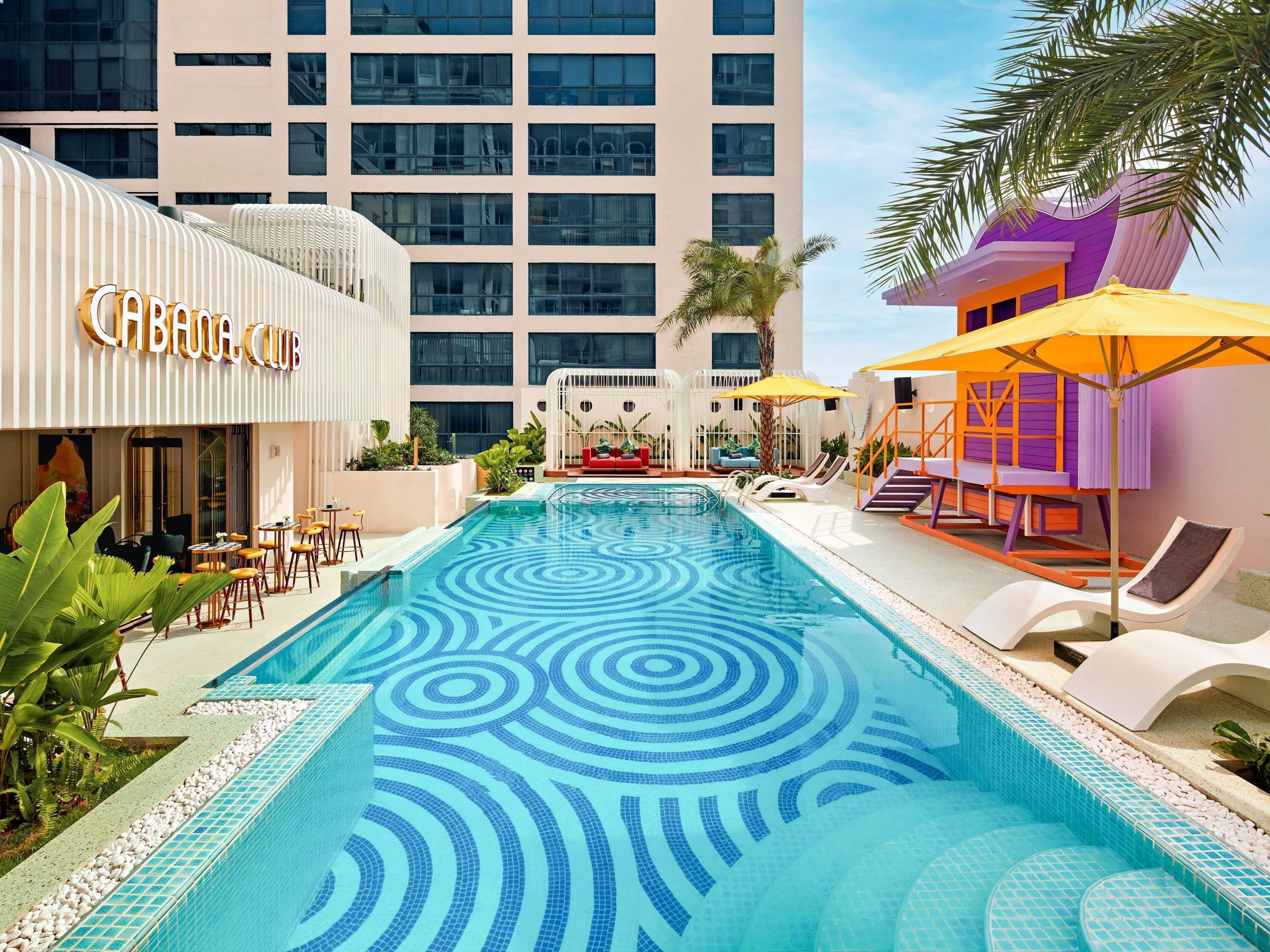 A swimming pool with a geometric tile pattern, surrounded by lounge chairs and umbrellas, with a building labeled 'CATHOLIC CLUB' in the background.