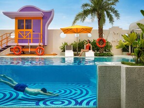 Una piscina con una torre de salvavidas, sillas de descanso y palmeras.