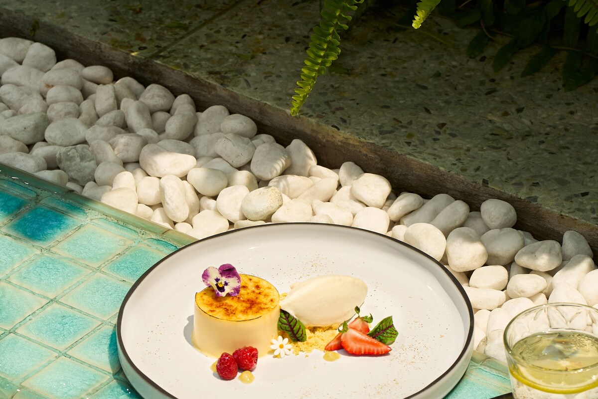 Un dessert présenté sur une assiette, placé au bord d’une piscine, entouré de petits galets blancs et de verdure.