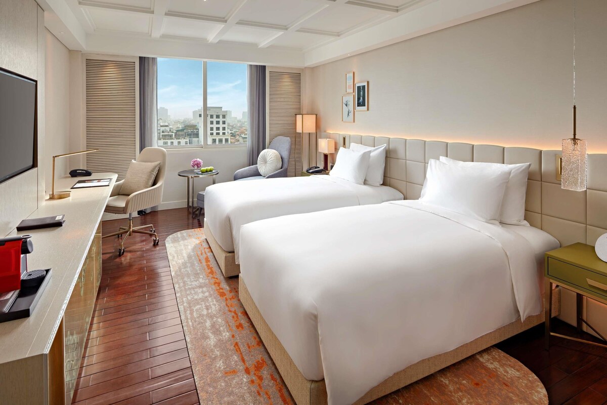 Ein Hotelzimmer mit zwei Betten, einem Schreibtisch, einem Stuhl, einem Fernseher und einem Fenster mit Blick auf die Stadt.