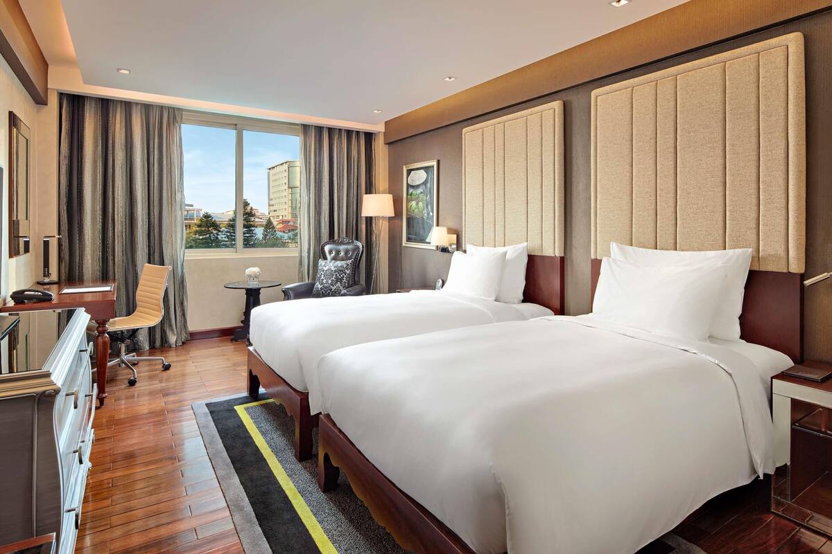Ein Hotelzimmer mit zwei Betten, einem Schreibtisch, einem Stuhl, einem kleinen Tisch und Blick auf die Stadt.