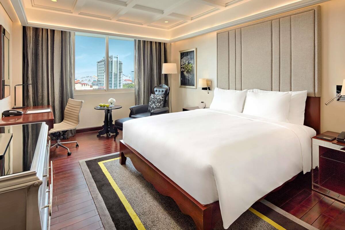 Ein Hotelzimmer mit einem großen Bett, einem Schreibtisch, einem Sessel, einem kleinen Tisch und Blick auf die Stadt.