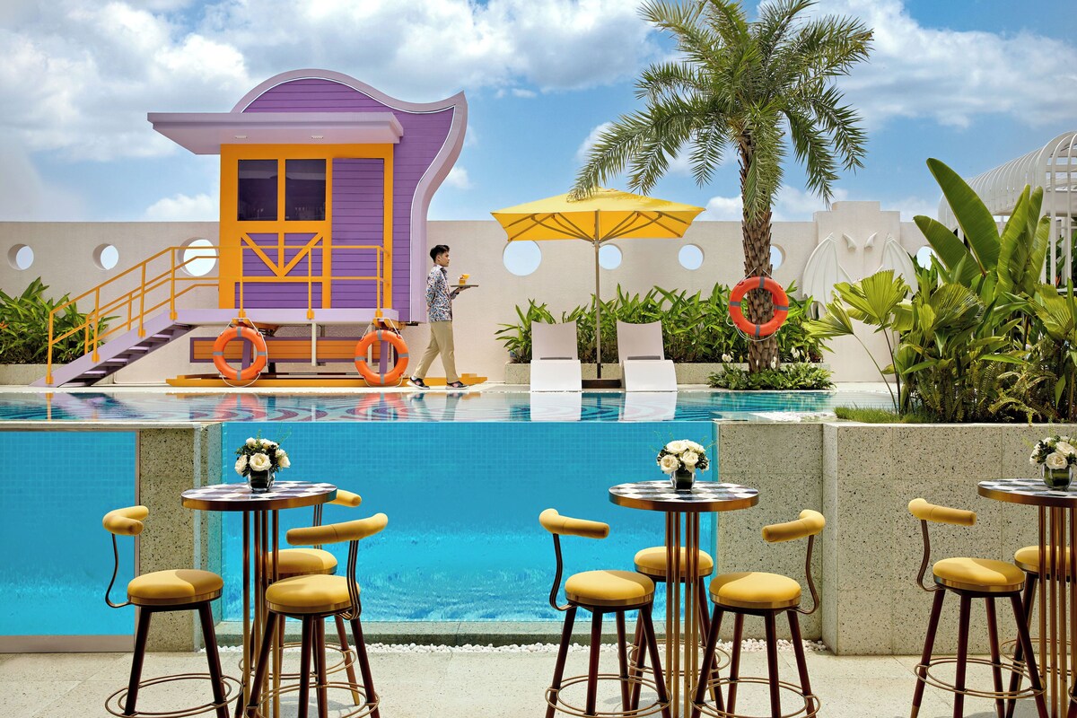 Un espace bar aménagé au bord de la piscine, avec plusieurs tables et chaises, une bouée de sauvetage et une tour de surveillance pour les sauveteurs.