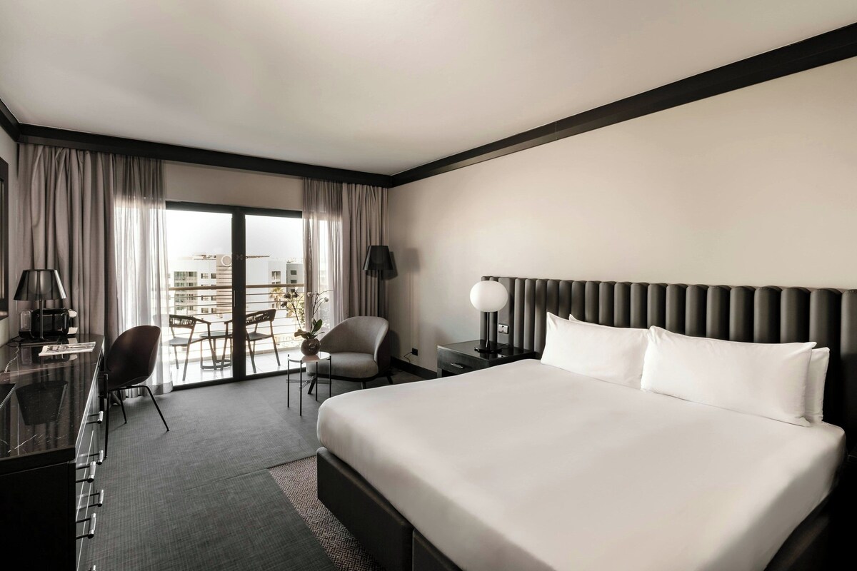 A hotel room with a large bed, a desk with a lamp, a chair, a small table with a plant, and a view of the city through the window.