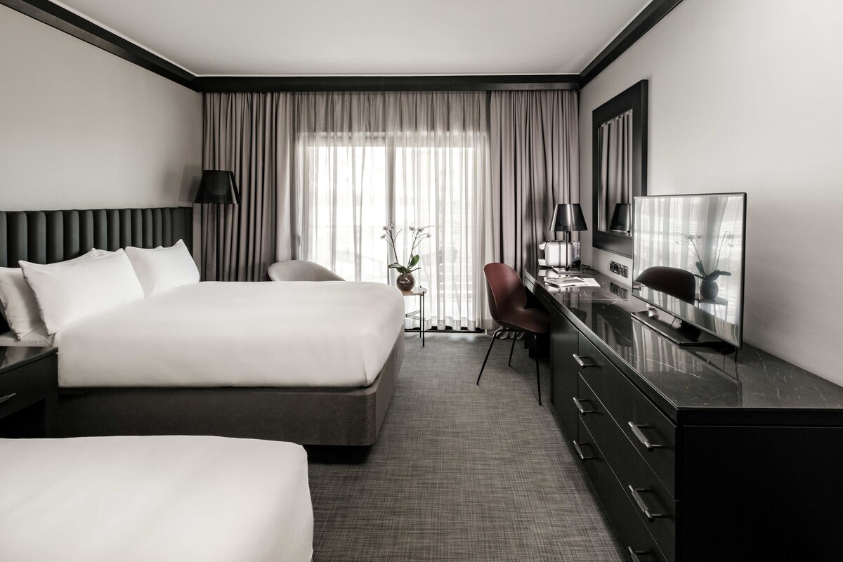 A hotel room with a large bed, a desk with a TV, a chair, and a small table with a vase of flowers.
