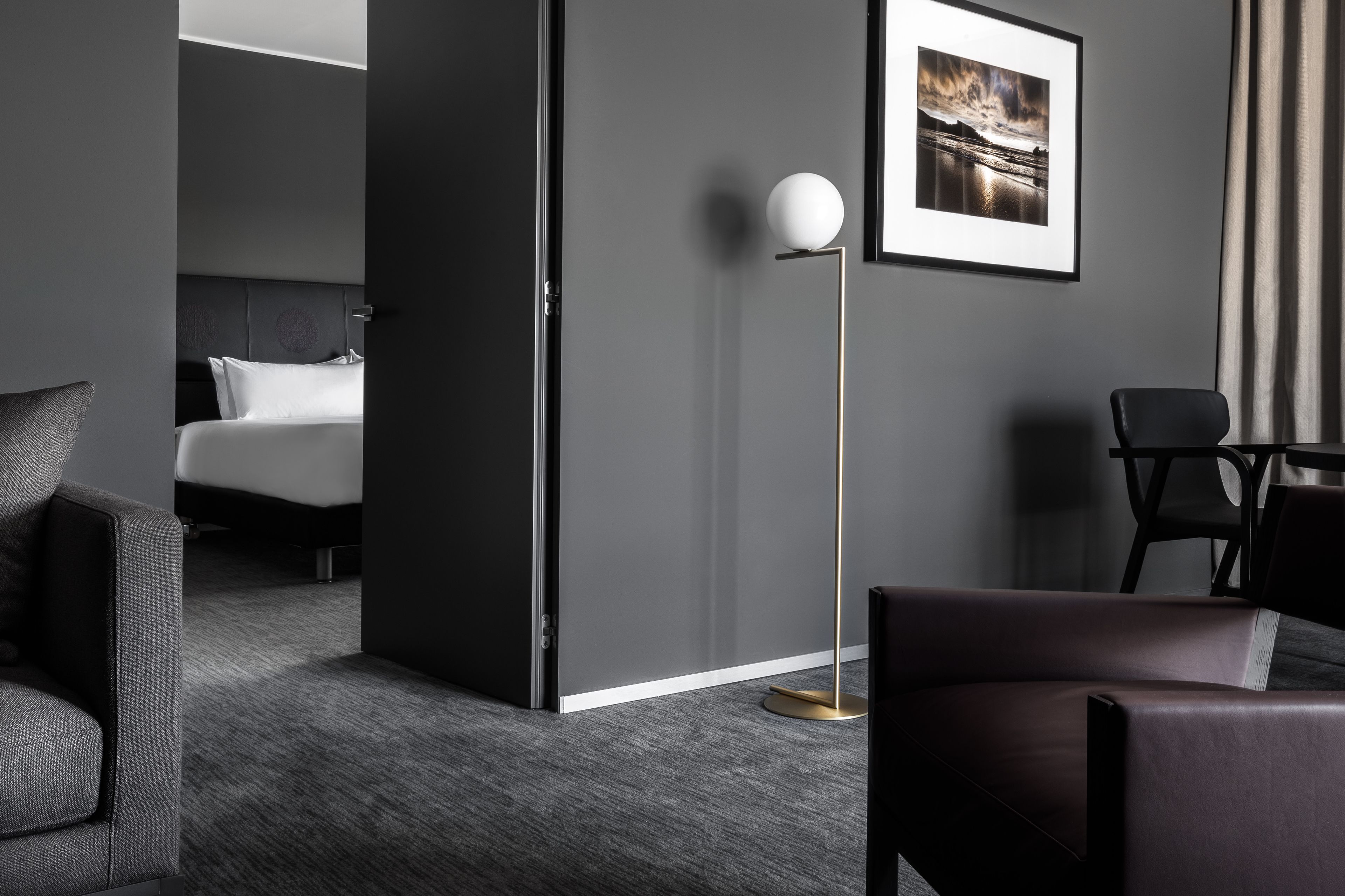 A modern hotel room with a dark grey wall, a framed landscape photo, a standing floor lamp, a bed with white linens, and a dark leather armchair.