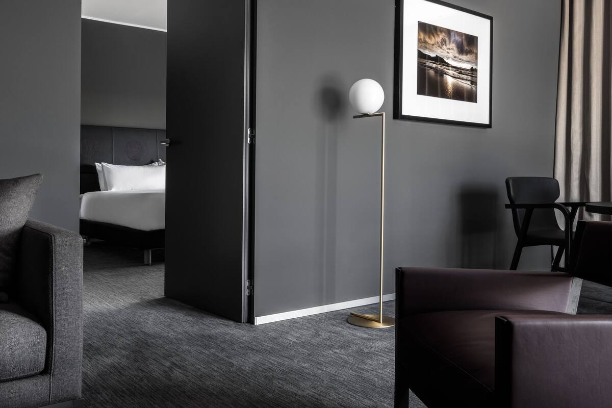 A modern hotel room with a dark grey wall, a framed landscape photo, a standing floor lamp, a bed with white linens, and a dark leather armchair.