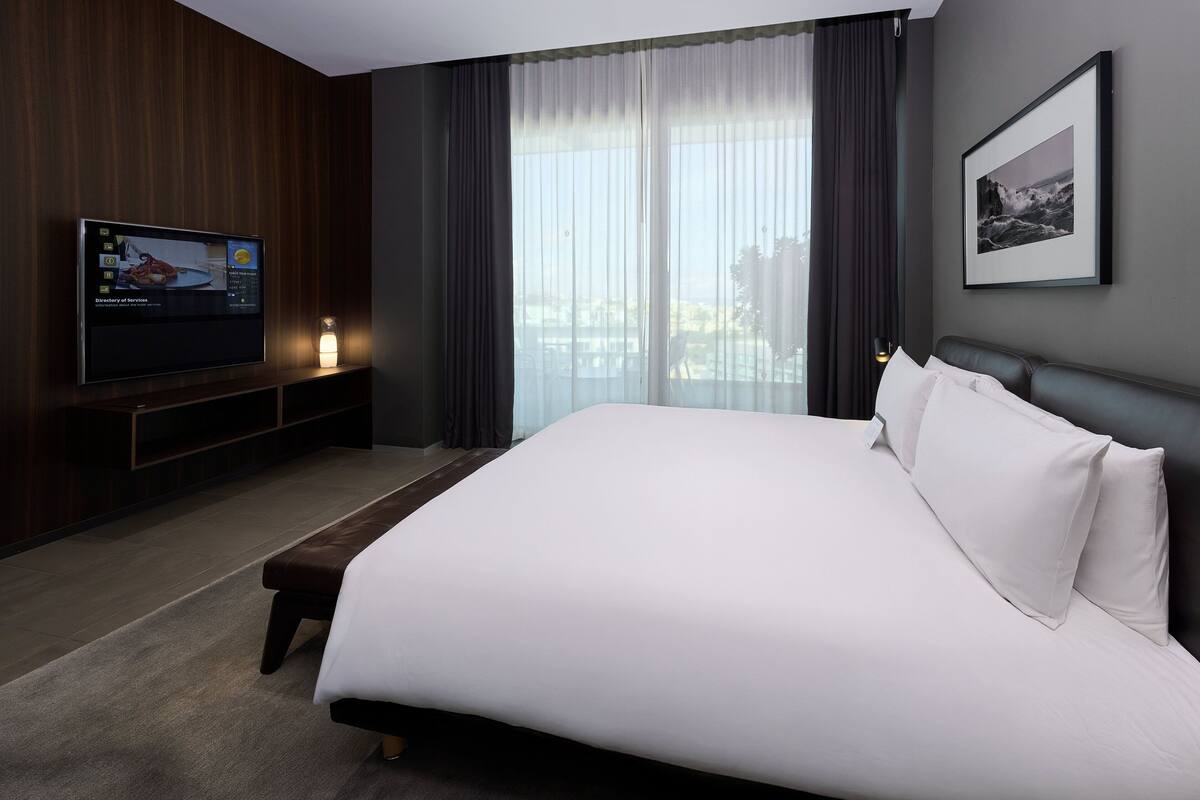A modern hotel room with a large bed, a flat-screen TV, a bedside table, and a large window with curtains.