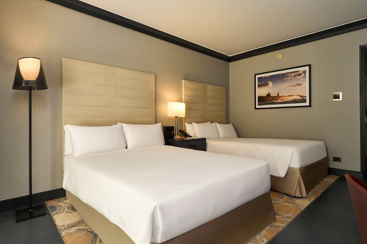 Quarto de hotel com duas camas, uma luminária de pé, mesa de cabeceira e um quadro na parede.