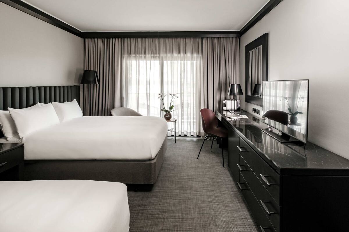 Quarto de hotel com uma cama grande, uma escrivaninha com televisão, uma cadeira e uma mesinha com um vaso de flores.